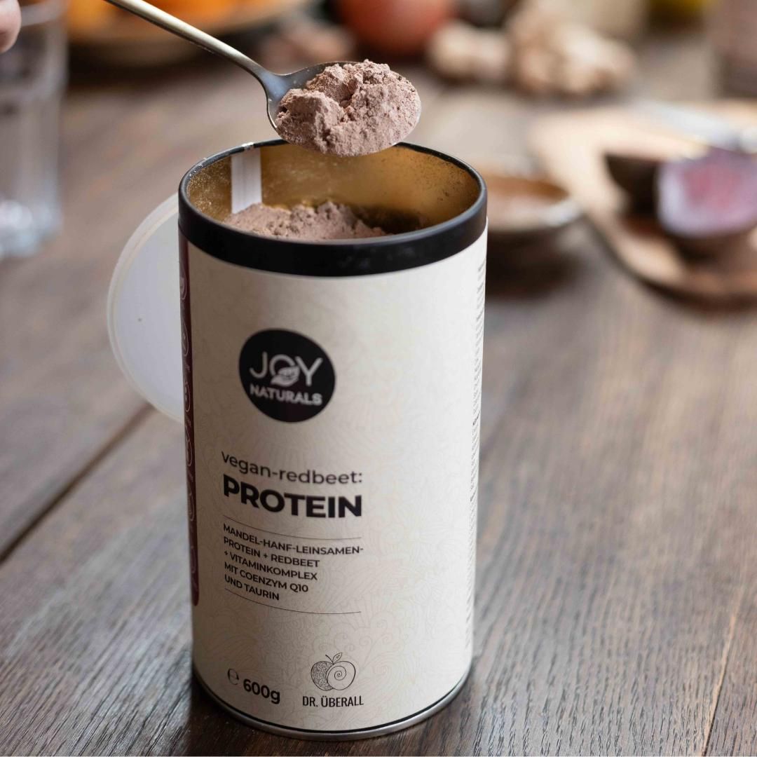 Beige Dose mit JOY NATURALS vegan-redbeet:PROTEIN. Löffel mit Pulver. Aufschrift: Protein, 600g.