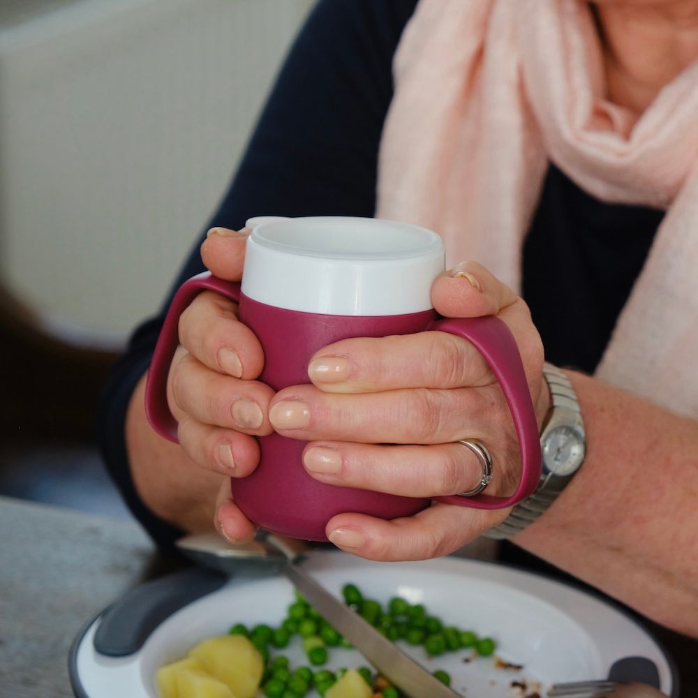 Person hält einen burgunderfarbenen Becher mit zwei Griffen. Der Becher hat einen weißen Deckel. Auf dem Tisch ein Teller mit Essen.