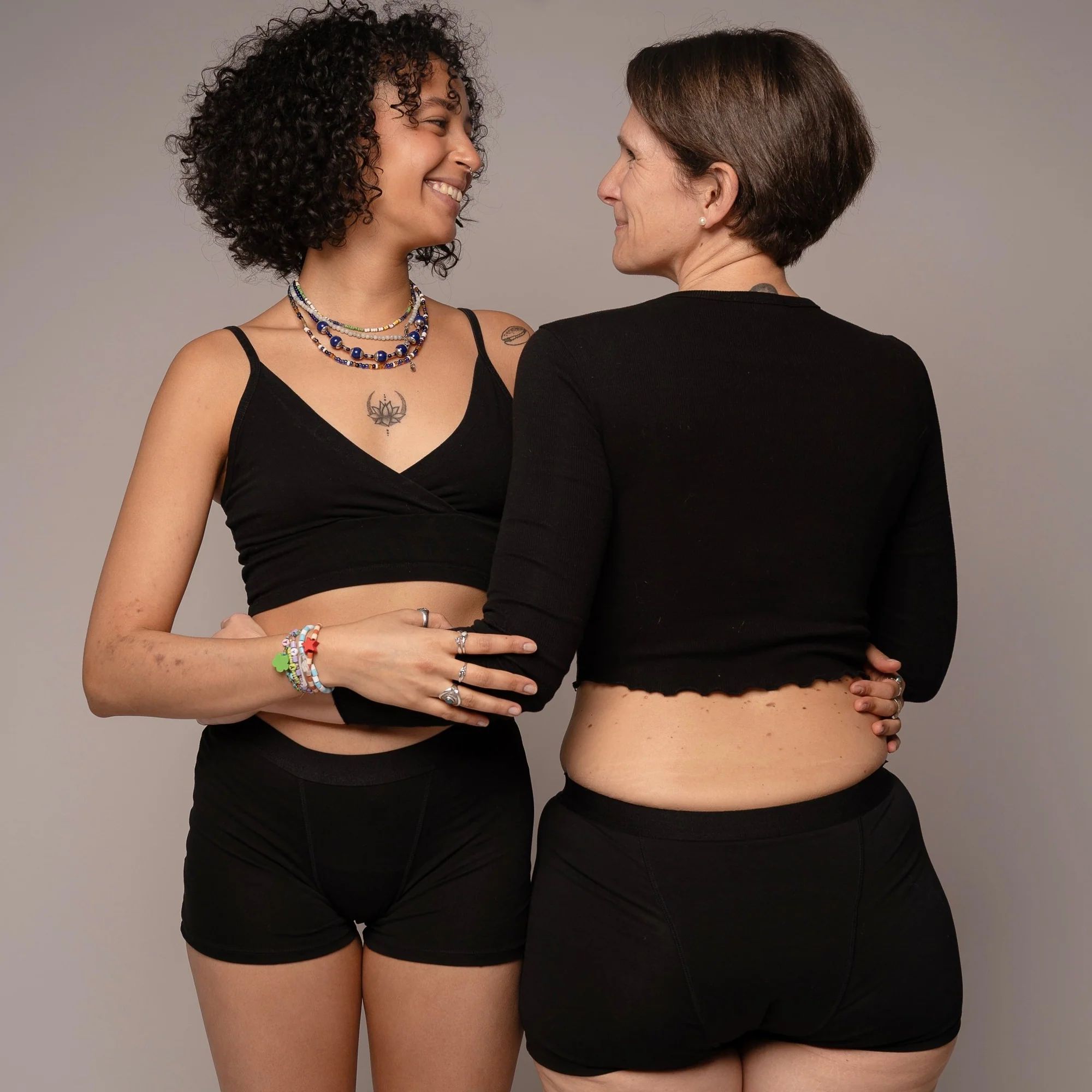 Zwei Personen umarmen sich. Beide tragen schwarze Tops und kurze Unterhosen. Person lächelt.