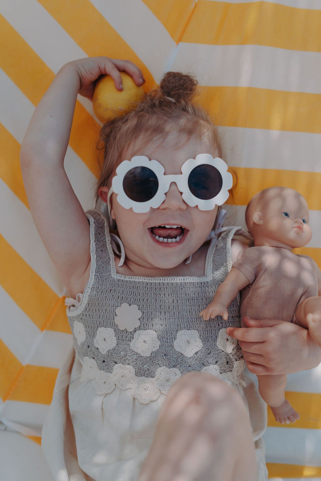 Kind mit Sonnenbrille und Puppe. Auf dem Kleid sind blütenförmige Sticker angebracht.