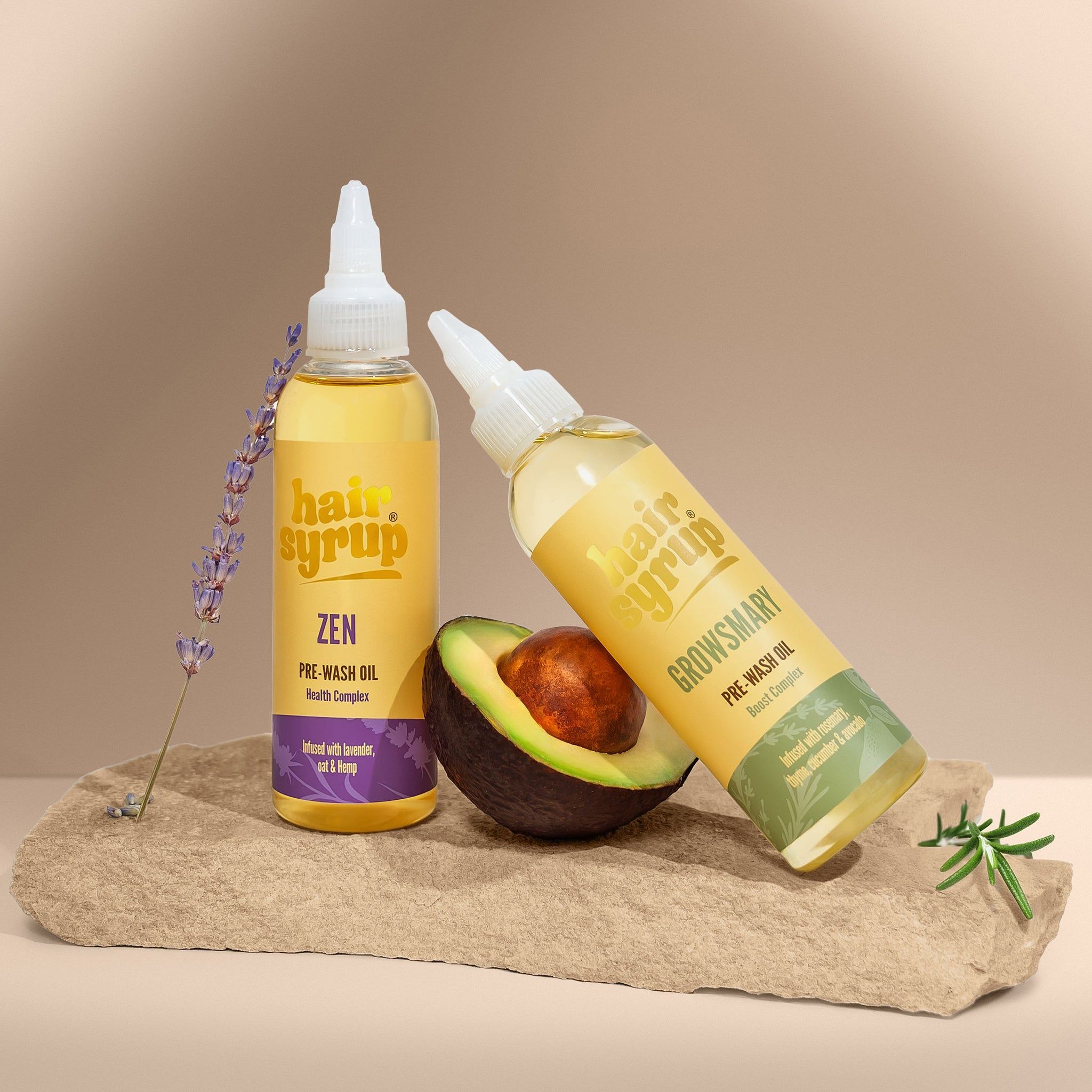 Zwei Flaschen mit gelber Flüssigkeit, Etiketten mit Text. Eine Flasche lehnt an einer Avocado. Lavendel und Stein im Hintergrund.