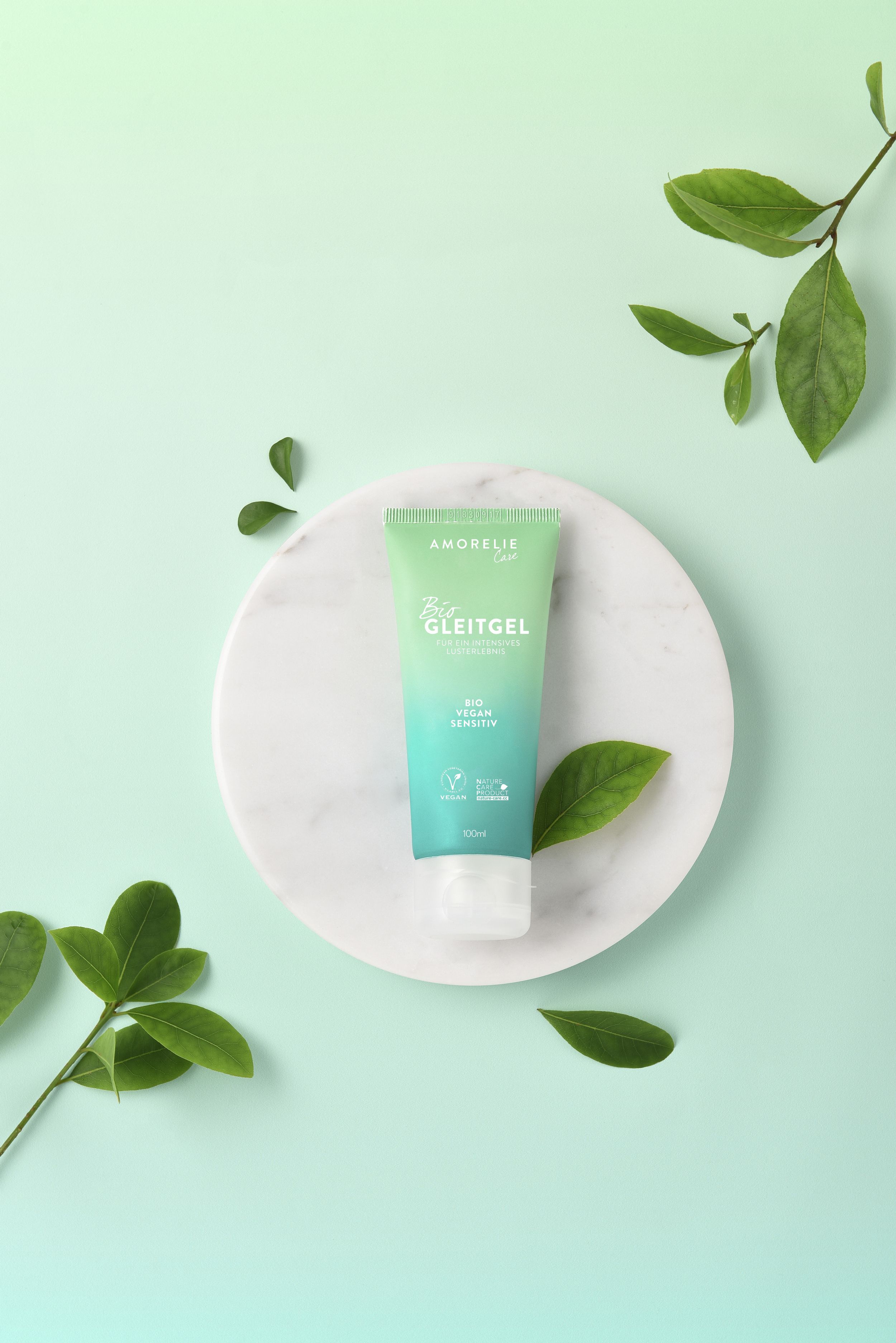 Tube vert sur marbre blanc. Inscription: AMORELIE Care, Bio Gleitgel, Bio Vegan Sensitiv. Label Vegan. Feuilles vertes.