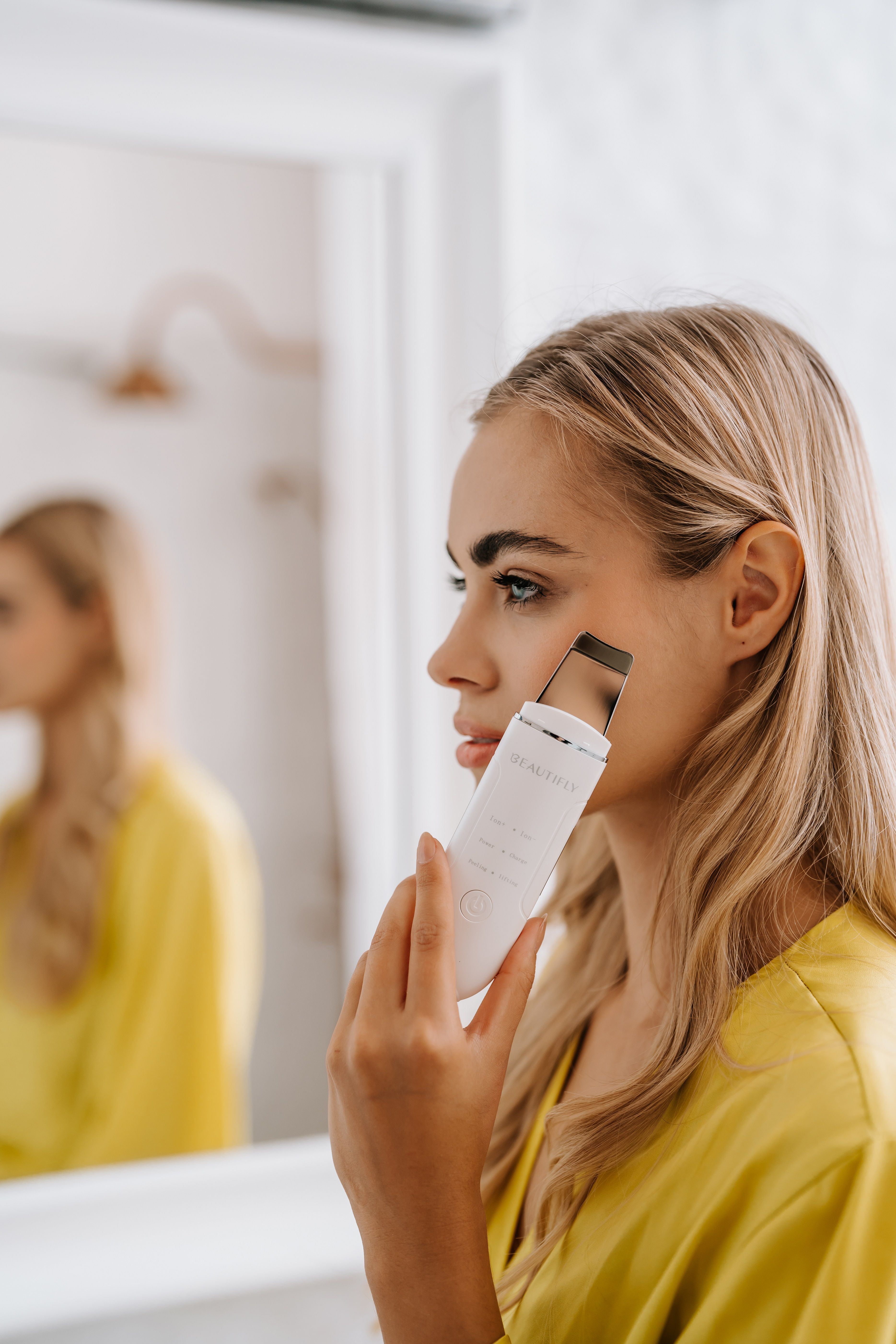 Frau hält ein weißes BEAUTIFLY B-Scrub Young Ultraschallpeelinggerät. Spiegel im Hintergrund. Gelbes Oberteil.