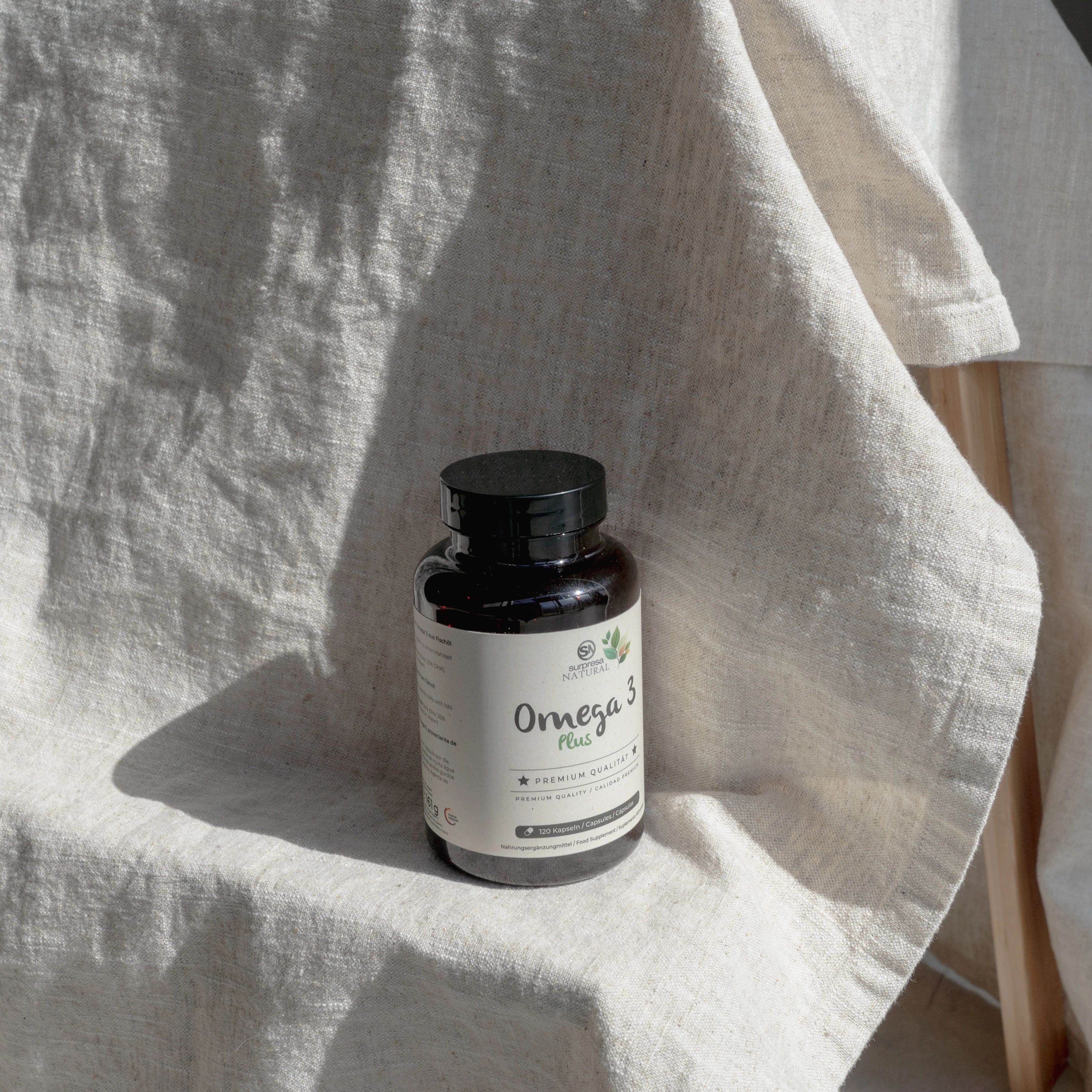 Braune Flasche mit schwarzen Deckel. Aufschrift: Omega 3 Plus. Steht auf hellem Stoff. Premium Qualität.