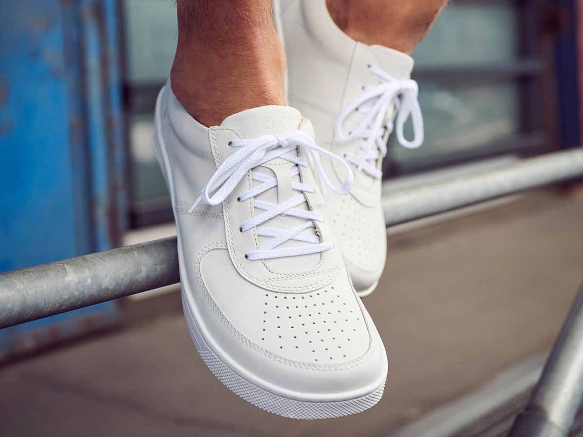 Weiße Lederschnürschuhe, breit, an Füßen. Perforiertes Vorderteil, weiße Schnürsenkel. Profilsohle. Füße sitzen auf Metallstange.