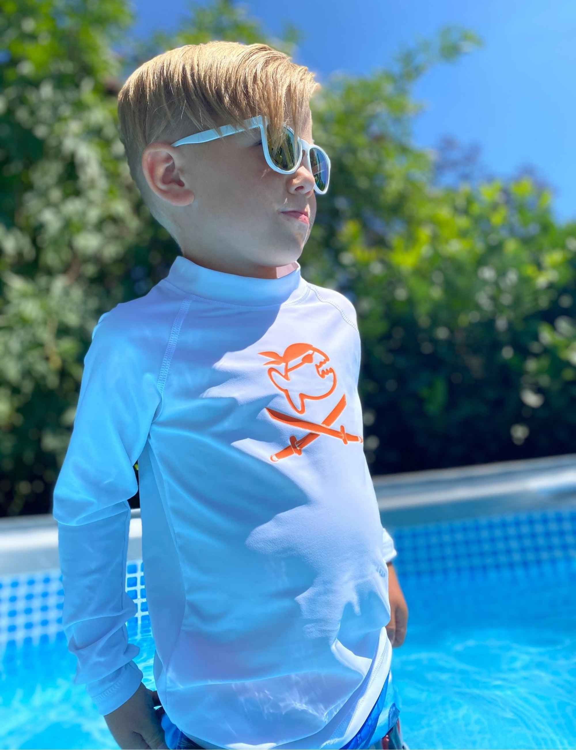 Kind mit weißem iQ-UV Sonnenschutz Shirt Jolly Kids am Pool. Langarm, hoher Kragen, orangefarbenes Logo. Trägt Sonnenbrille.