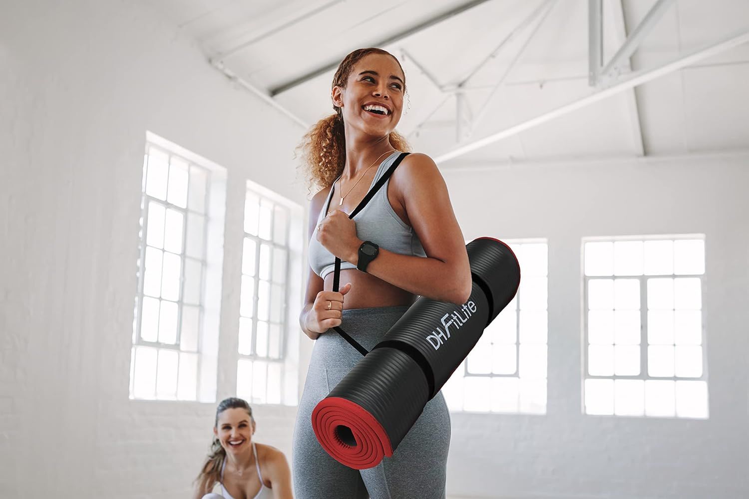 Frau trägt gerollte Trainingsmatte mit DH FitLife Logo. Schwarze Matte mit rotem Rand. Im Fitnessstudio.