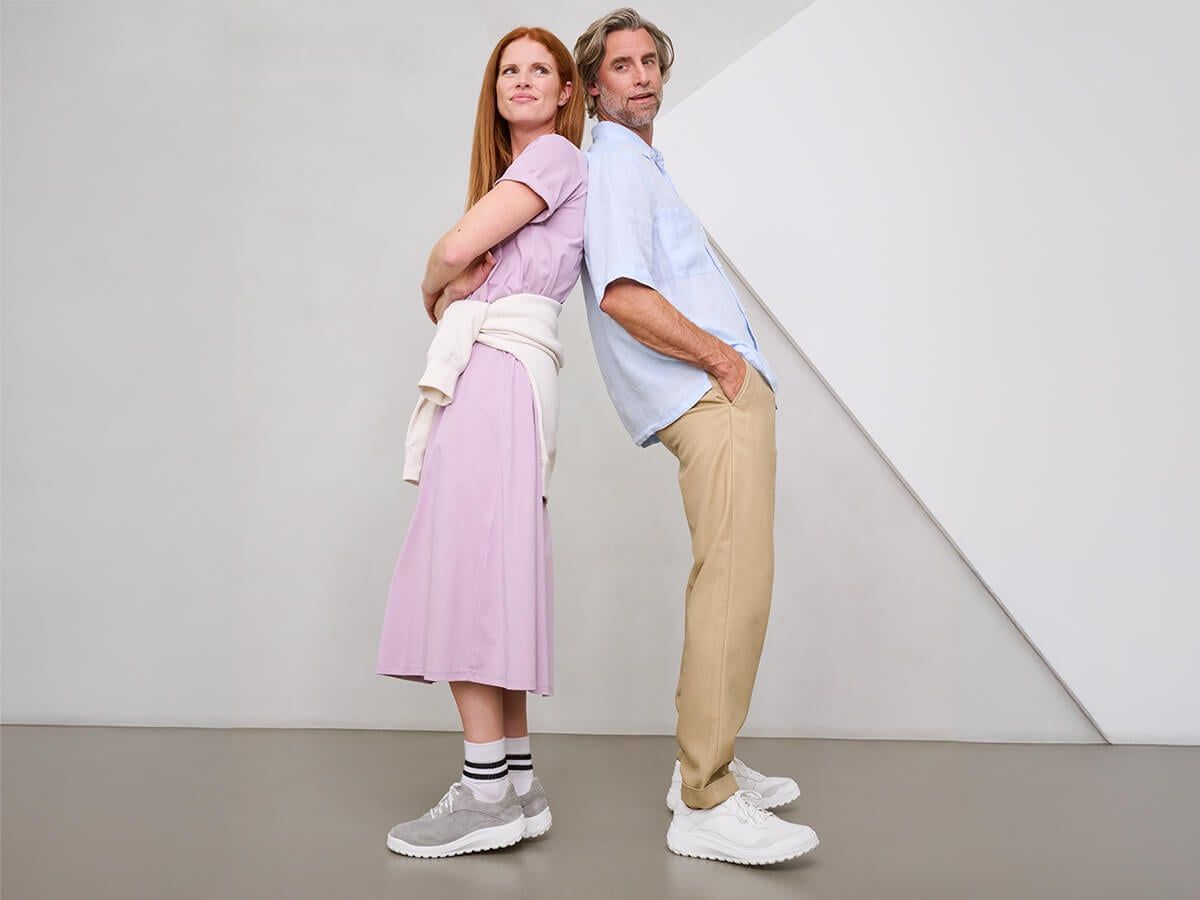 Ein Mann und eine Frau stehen Rücken an Rücken. Beide tragen helle Kleidung und Schuhe. Die Frau hat rote Haare.