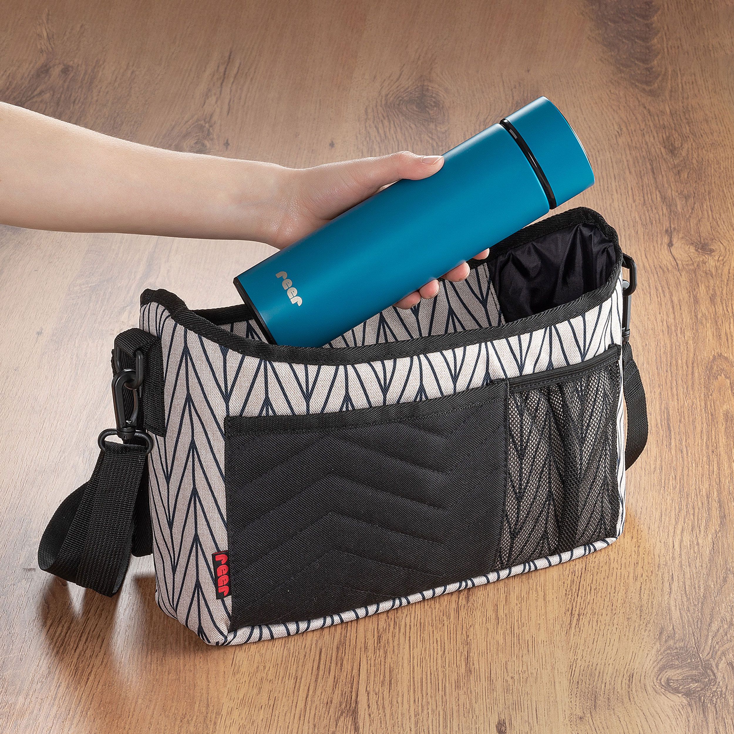 Blaue Isolierflasche wird in eine Wickeltasche gelegt. Markenlogo sichtbar. Auf Holzuntergrund.