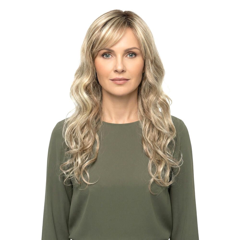 Blond gewellte Perücke mit Pony, frontal. Die Frau trägt ein grünes Oberteil. Hintergrund weiß.