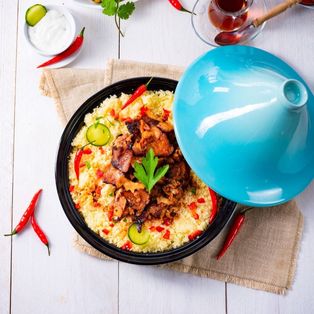 Tajine-Gericht mit Fleisch, Gemüse und Couscous. Blaue Tajine-Haube. Rote Chilischoten und Limettenscheiben als Garnitur.