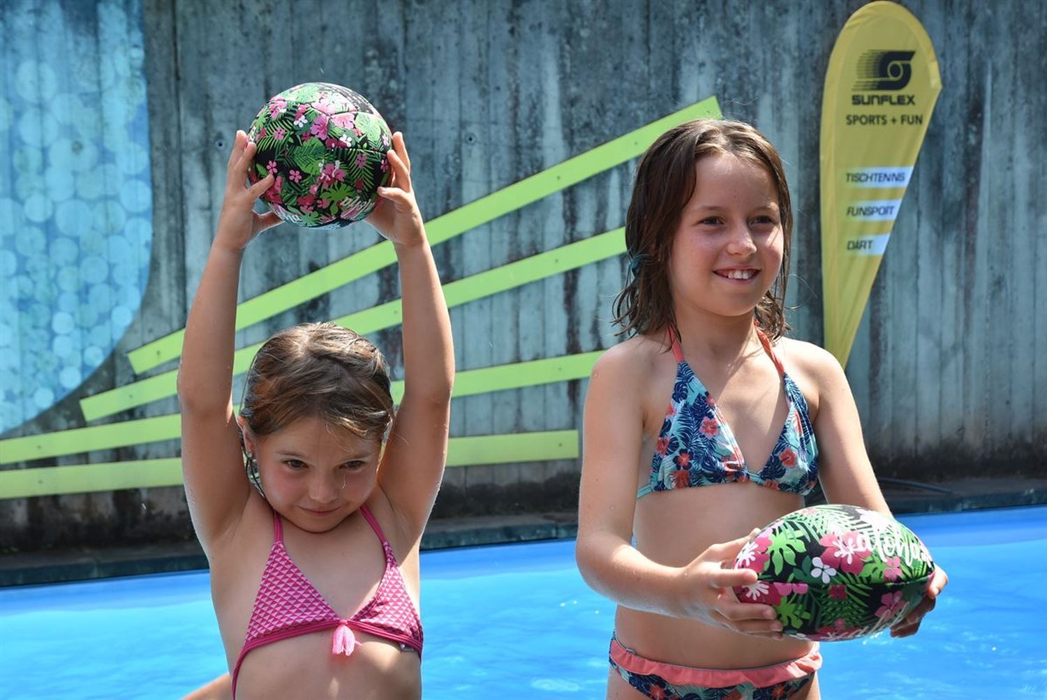Zwei Mädchen im Wasser spielen mit Bällen mit tropischem Blumenmuster. Beide halten einen Ball.