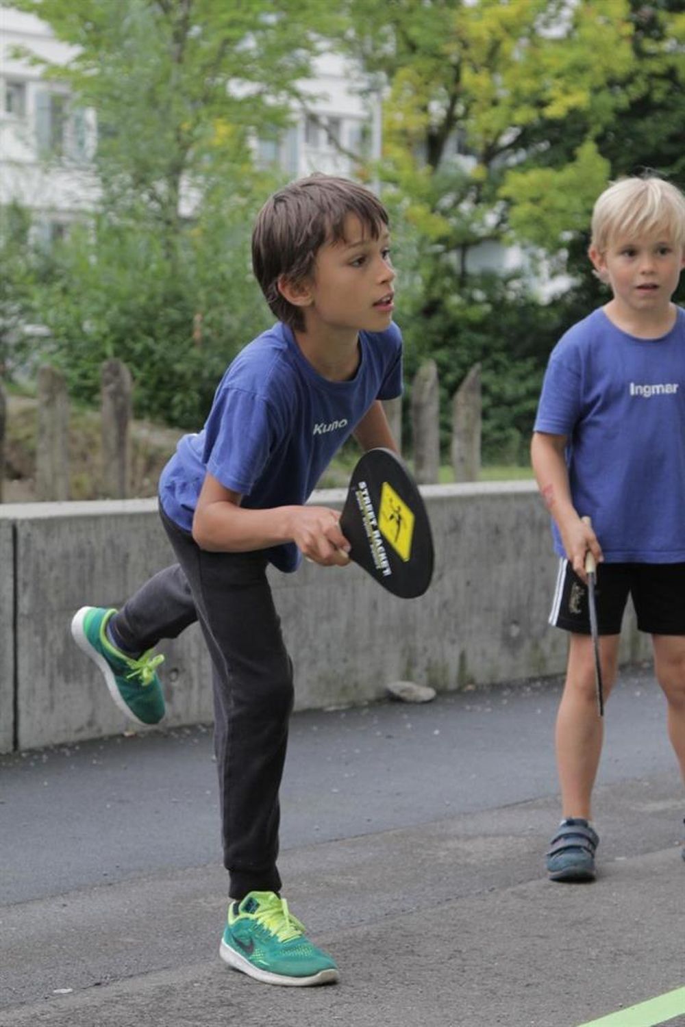 Junge spielt Street Racket. Er hält einen schwarzen Schläger mit gelbem Logo. Er trägt ein blaues T-Shirt und graue Hose.