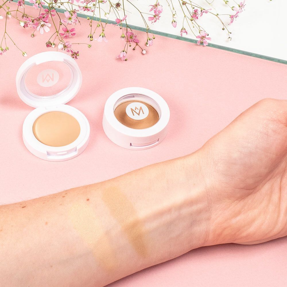 Zwei runde, weiße Concealer-Behälter. Geöffnet und geschlossen. Arm mit zwei Farbproben. Rosa Hintergrund.