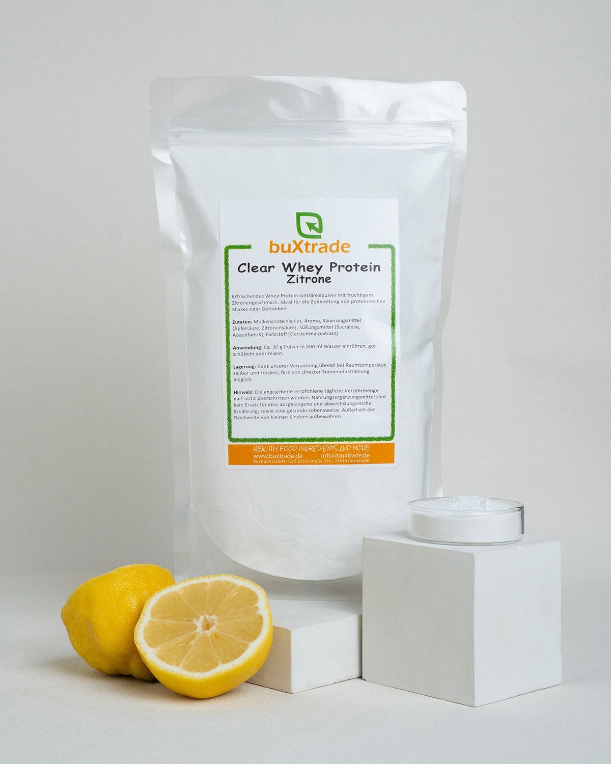Buxtrade Clear Whey Protein Zitrone in einem weißen Beutel, daneben Zitronen und ein kleiner Behälter mit Pulver.
