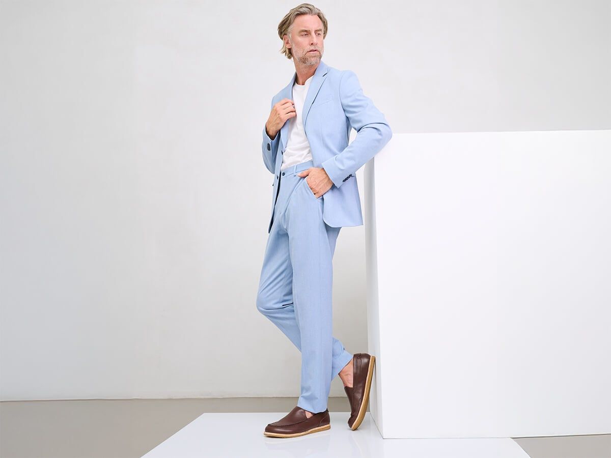 Mann in hellblauem Anzug und braunen Slippern. Steht an weißer Wand.