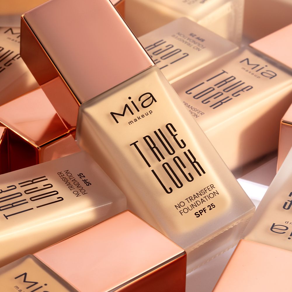 Diversi flaconi di fondotinta Mia Makeup True Lock. I flaconi sono in vetro con tappi color oro rosa. Le etichette riportano "True Lock" e "No Transfer Foundation SPF 25". I flaconi sono parzialmente sfocati sullo sfondo.