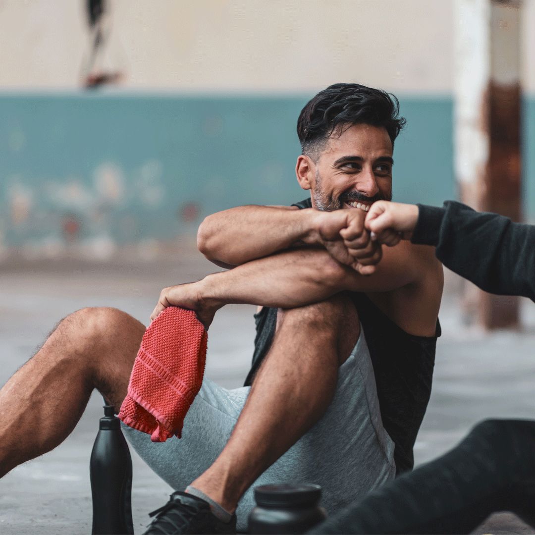 Mann sitzt auf dem Boden, gibt einem anderen Mann einen Fauststoß. Rotes Handtuch und schwarze Flasche daneben.