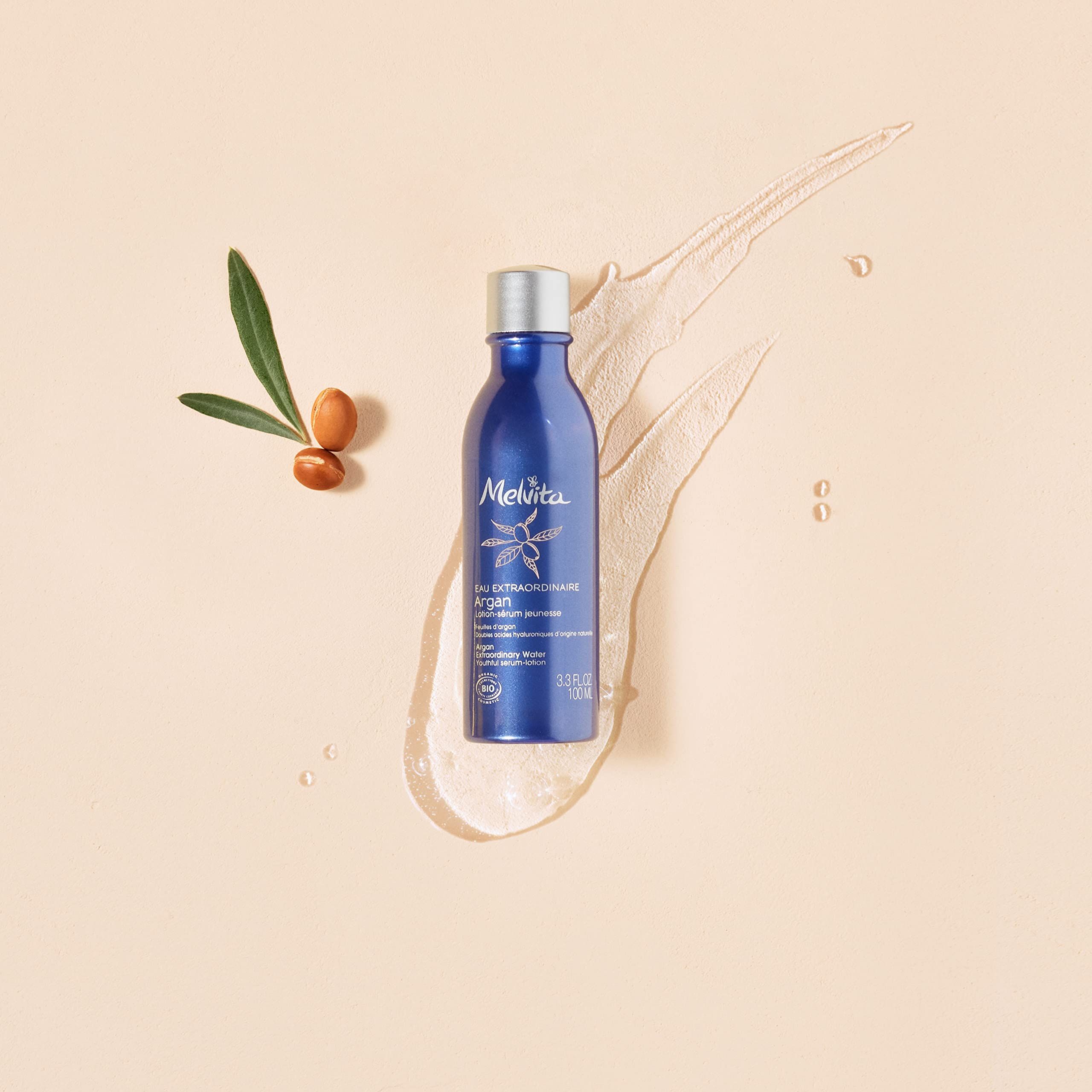 Blaue Flasche mit silbernem Deckel. Daneben Argan-Blatt und -Samen. Flüssigkeitsspritzer. Aufschrift Melvita.
