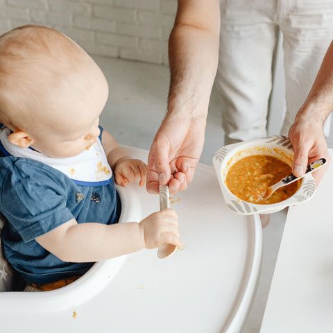 Baby im Hochstuhl, Löffel in der Hand, Person reicht Schüssel mit Brei.