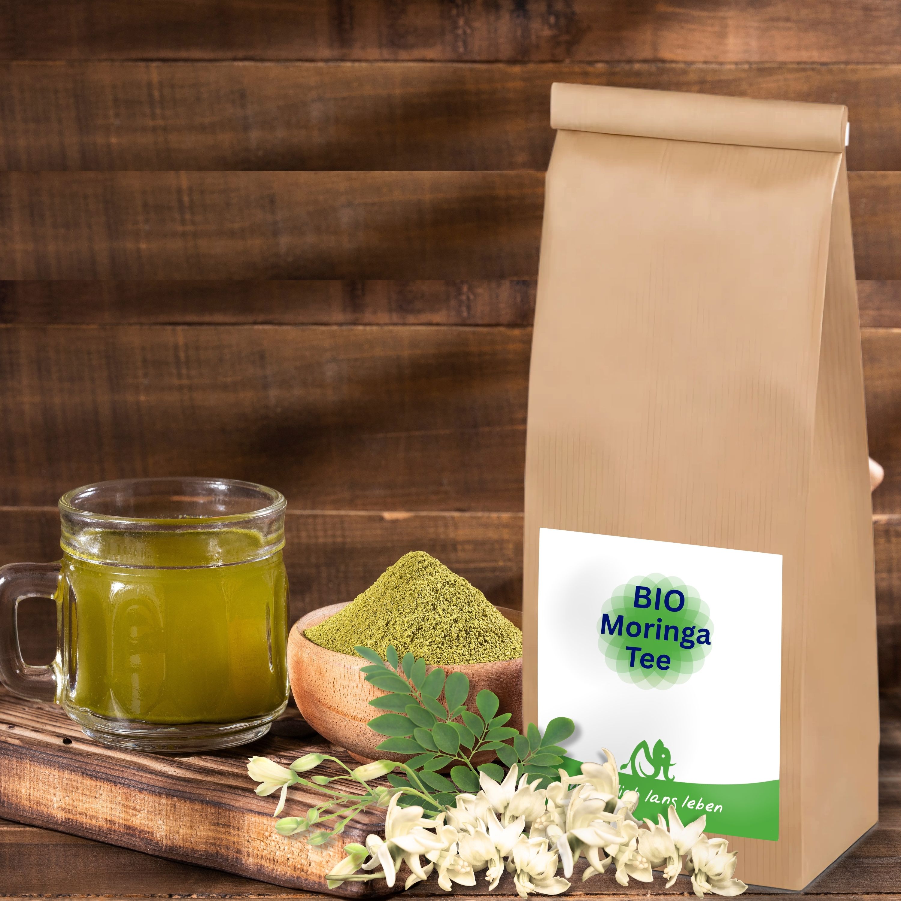 Braune Papiertüte mit Etikett, daneben Glas mit grünem Getränk, Holzschale mit Pulver, Blätter und Blüten. Etikett: BIO Moringa Tee.