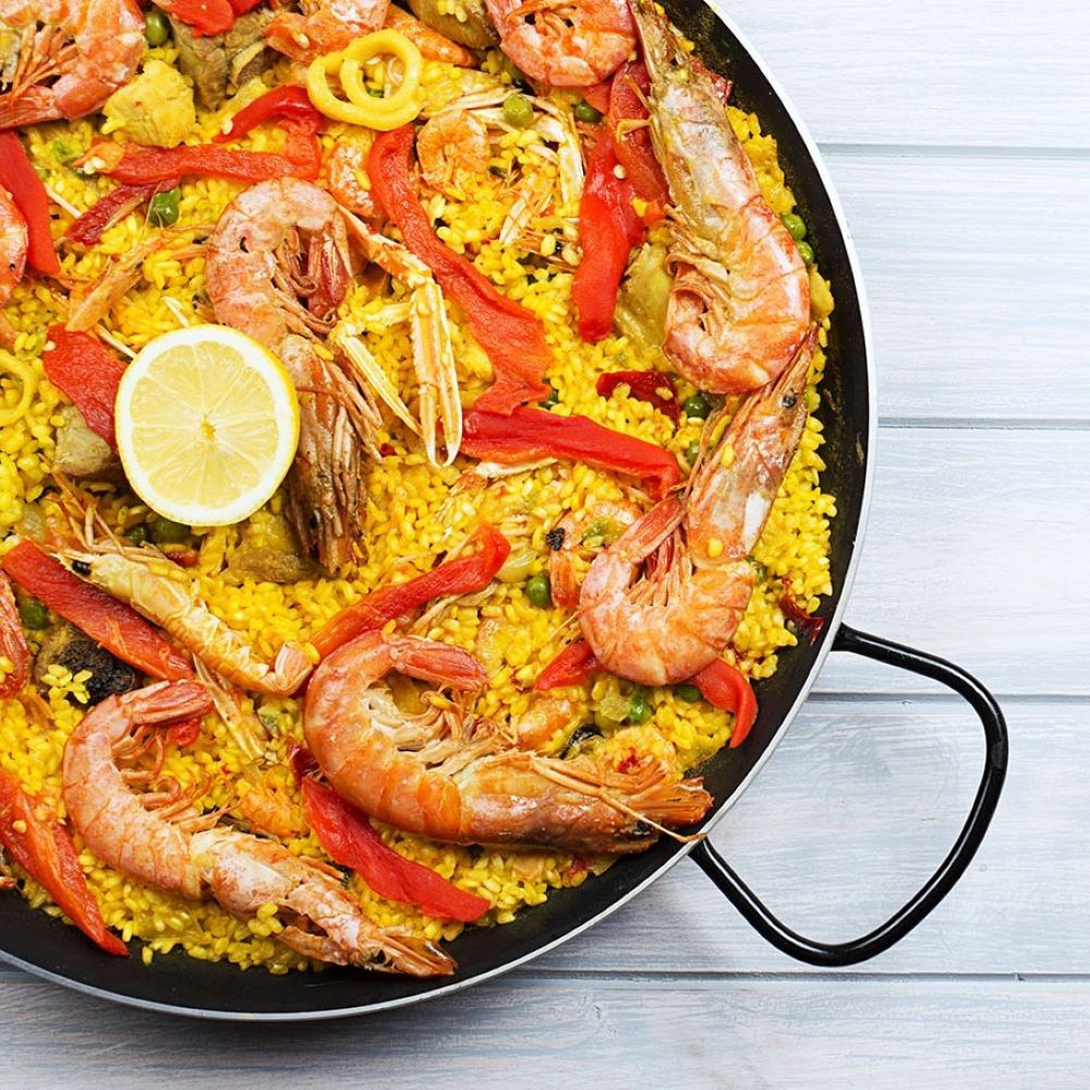 Paella mit Garnelen, Zitrone und Gemüse in einer schwarzen Pfanne. Auf hellem Holzuntergrund. Draufsicht.