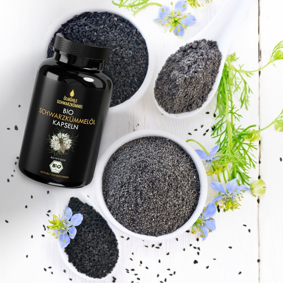 Schwarze Flasche mit BIO Schwarzkümmelöl Kapseln neben Schalen mit Samen und Pulver. Blüten und Löffel.