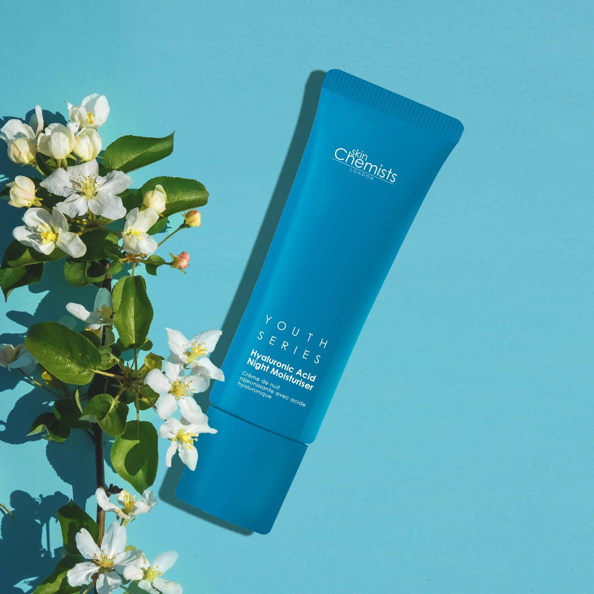 Blaue Tube mit Produktbezeichnung und Schriftzug. Marke: skinChemists. Produkt: Youth Series Hyaluronsäure Nacht-Feuchtigkeitscreme. Blumen.