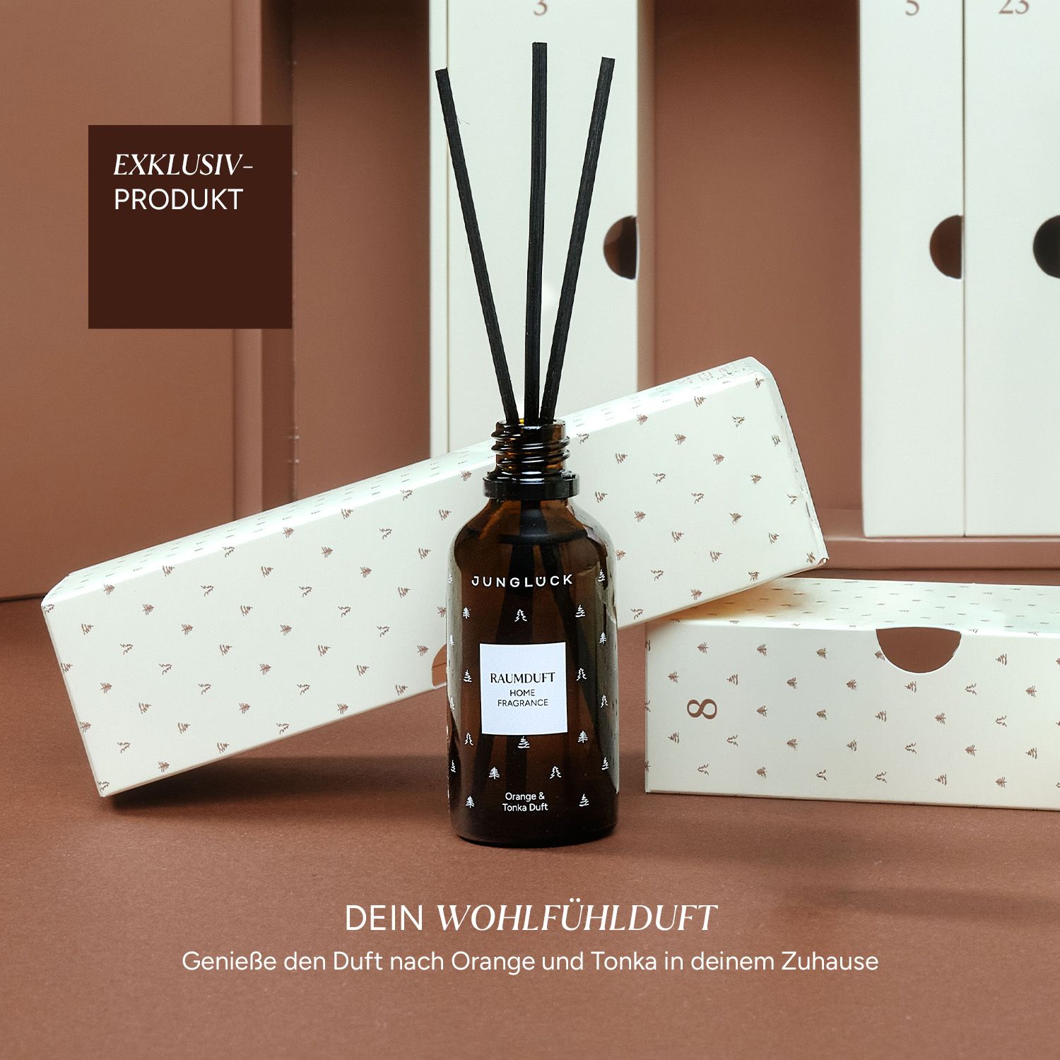 Braune Glasflasche mit schwarzen Stäbchen. Weiße Schachtel mit Dreiecksmuster. Text: JUNGLÜCK.