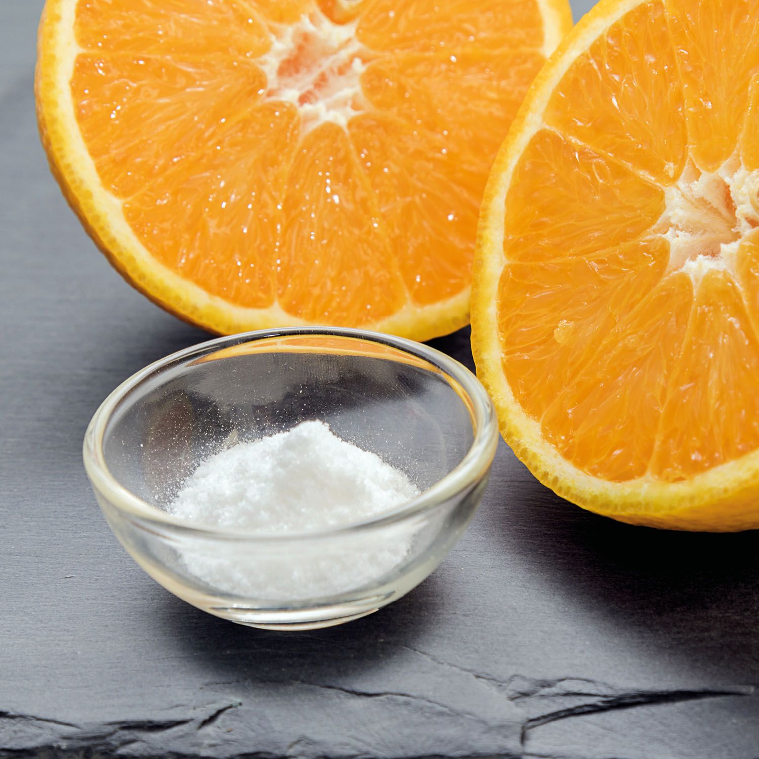 Kleine Glasschale mit weißem Pulver neben zwei halbierten Orangen auf dunklem Untergrund.