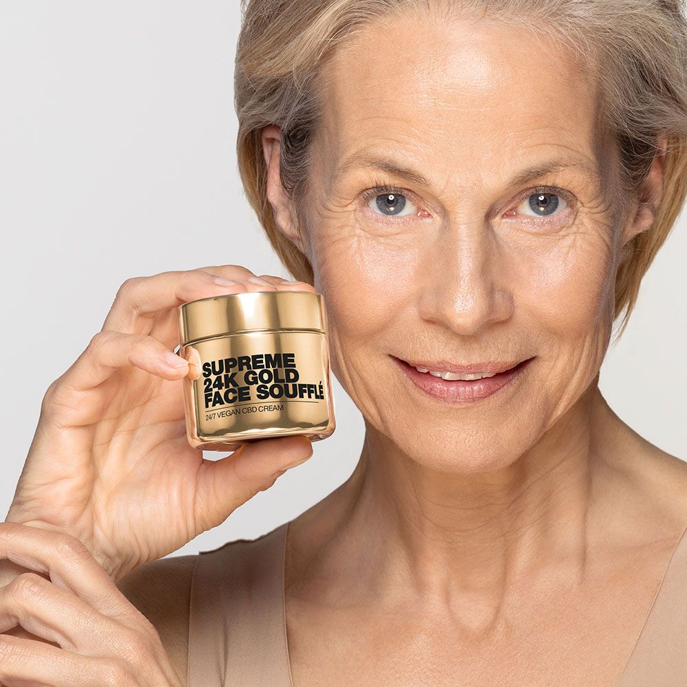 Ältere Frau hält goldfarbenen Tiegel. Text: Supreme 24K Gold Face Soufflé. 24/7 Vegan CBD Cream. Frau lächelt.