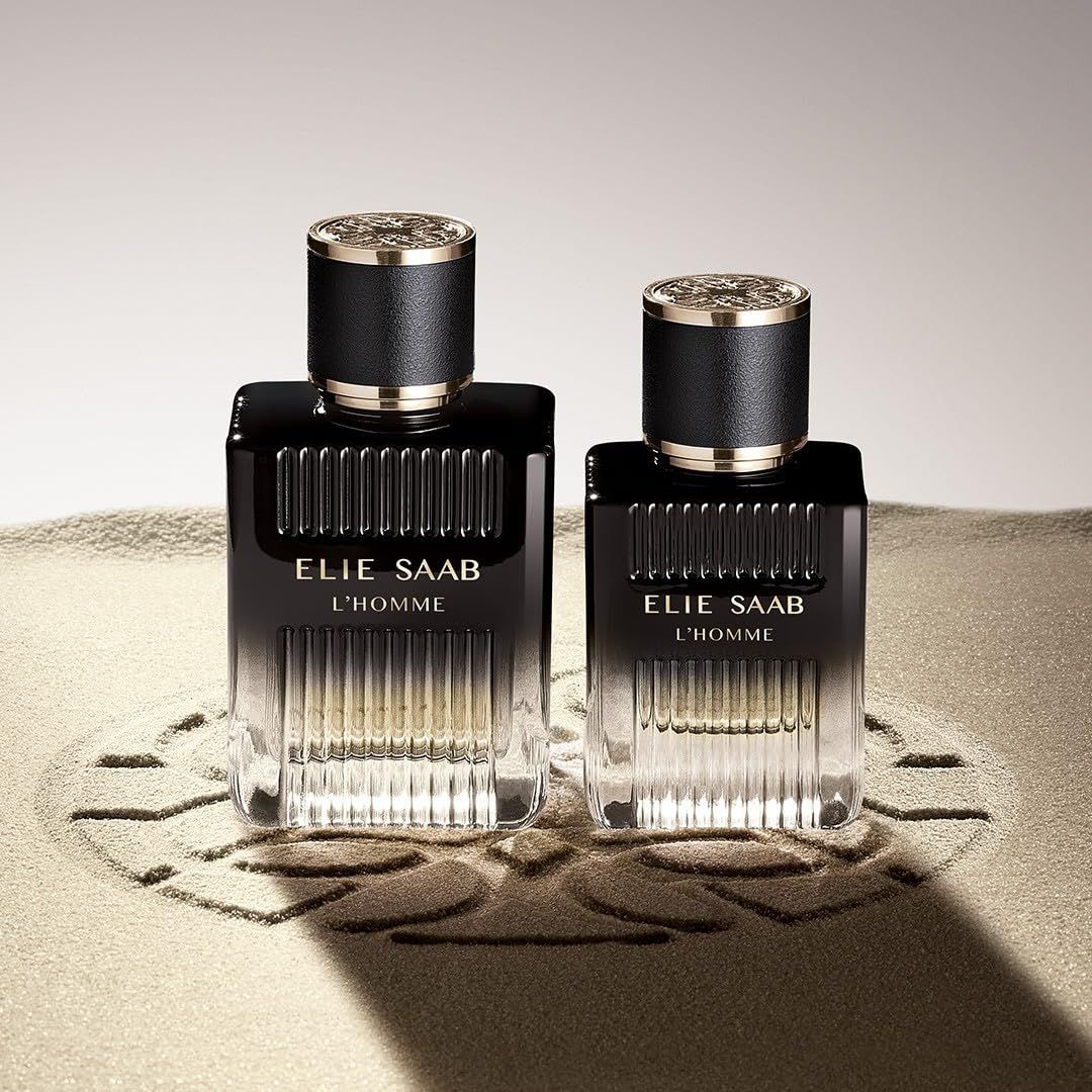 Zwei schwarze Flakons mit goldfarbenen Akzenten. Aufschrift: ELIE SAAB L'HOMME. Auf Sand platziert.