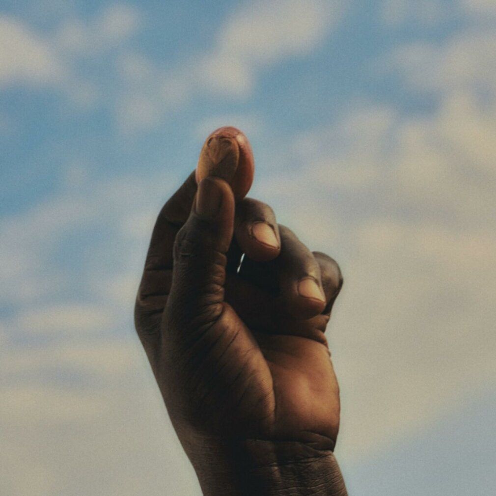 Nahaufnahme einer Hand, die eine Nuss hält, vor blauem Himmel mit Wolken. Die Haut ist dunkel.