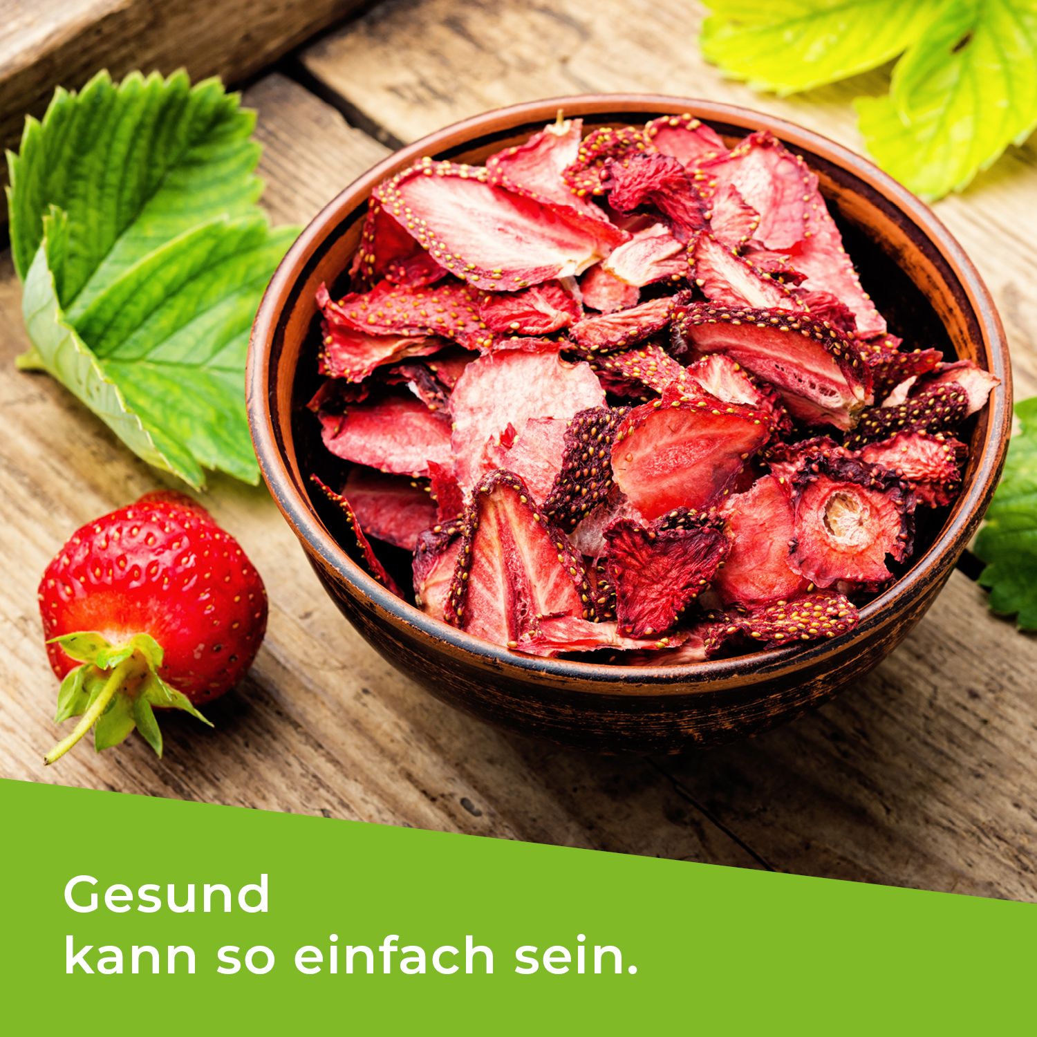 Schale mit gefriergetrockneten Erdbeerscheiben, daneben eine frische Erdbeere und Blätter. Text: Gesund kann so einfach sein.