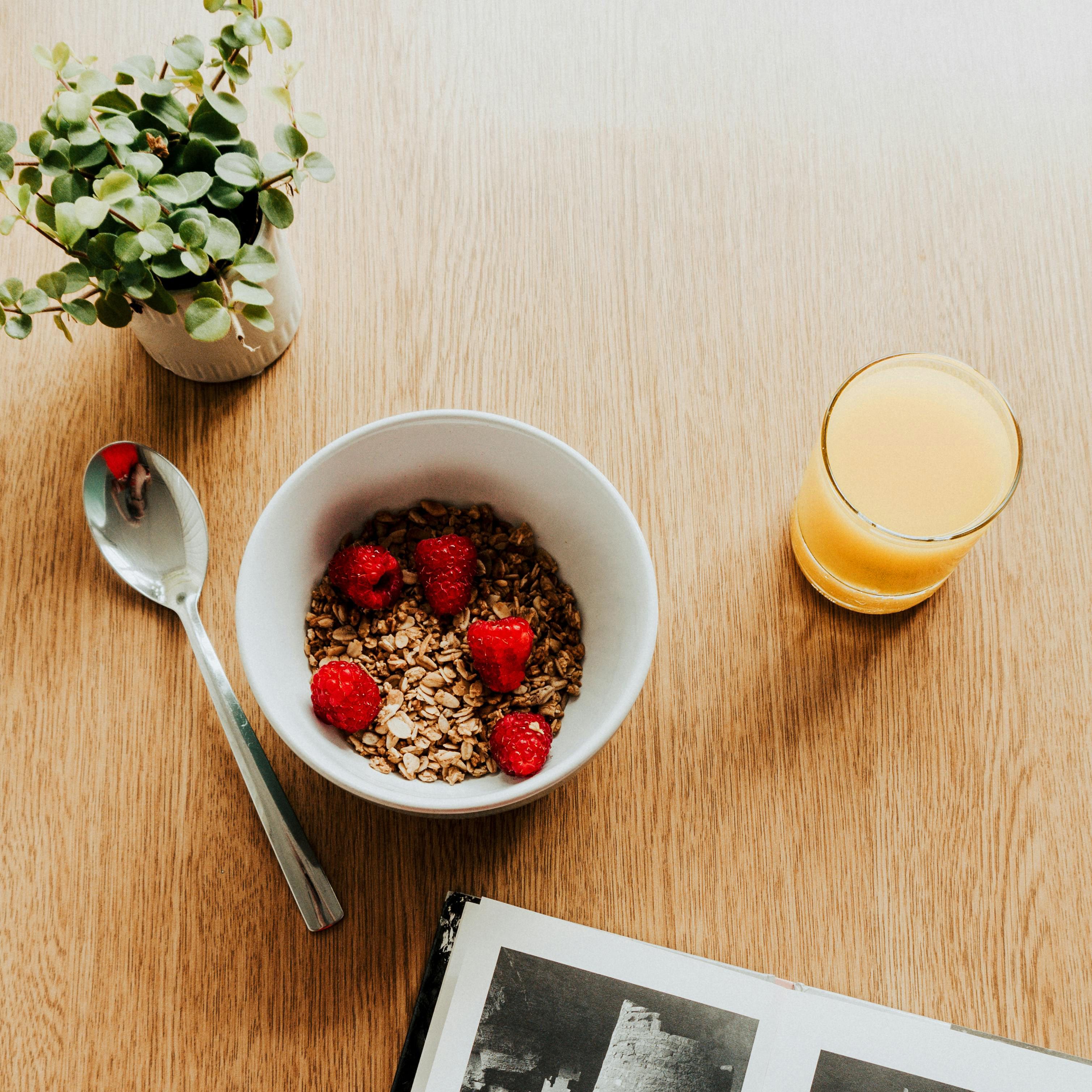 Draufsicht auf Holztisch mit Schüssel Müsli mit Himbeeren, Glas Saft, Löffel und Buch.