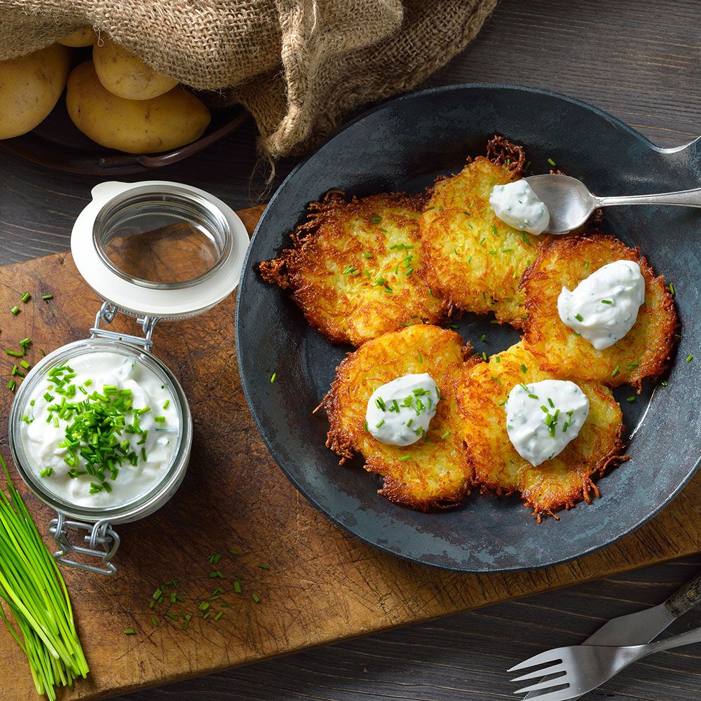 Gebratene Kartoffelpuffer mit Quark und Schnittlauch in einer Pfanne. Daneben ein Glas mit Quark und Schnittlauch. Holzbrett.