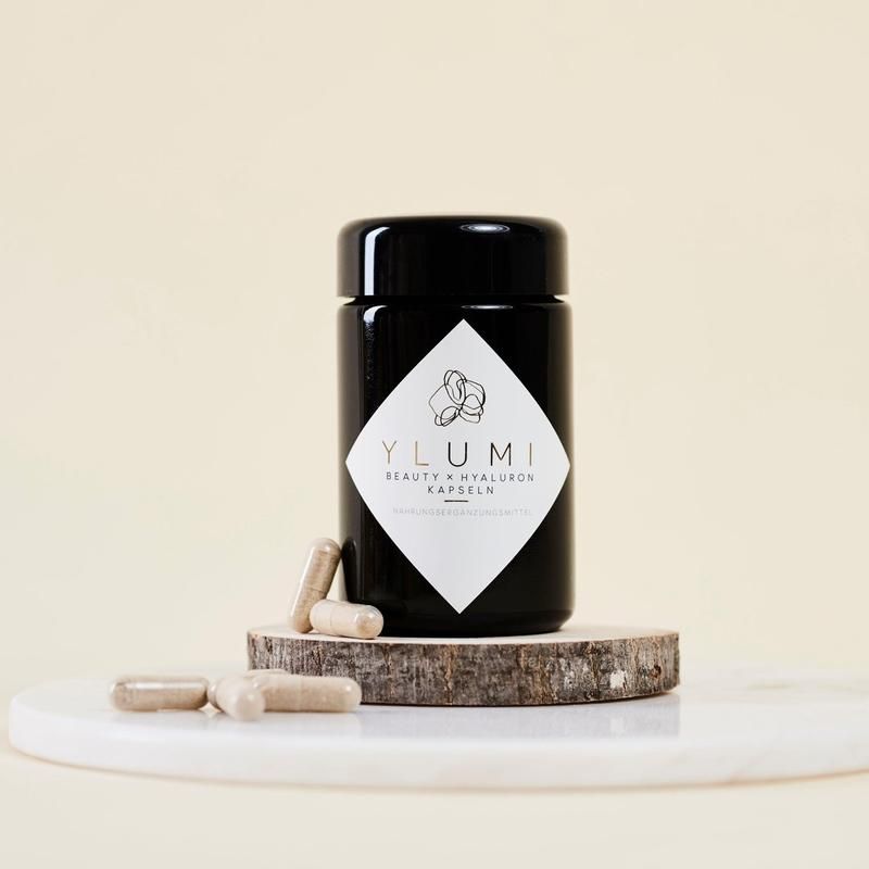 Schwarze Dose mit weißem Etikett auf Holzscheibe. Aufschrift: YLUMI BEAUTY x HYALURON KAPSELN. Daneben Kapseln.