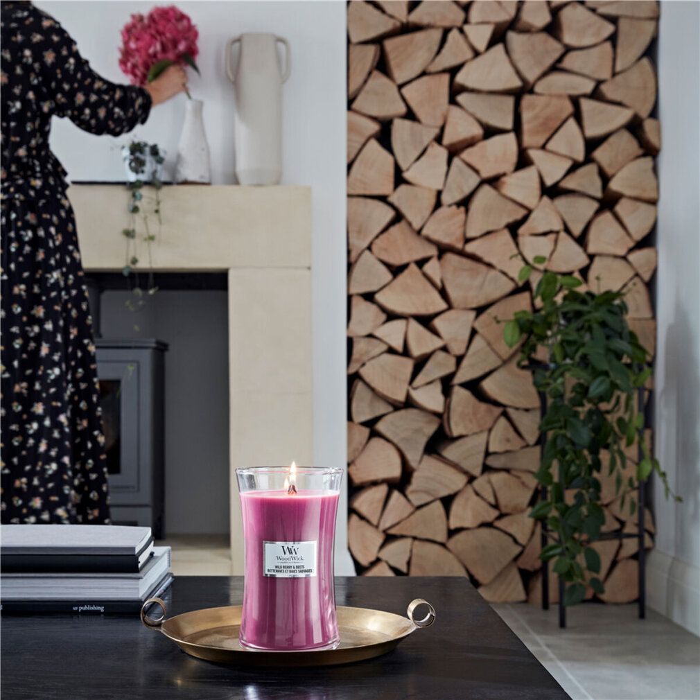Brennende Duftkerze mit Holzdocht auf Tablett. Frau mit Blumen im Hintergrund. Holzstapel.