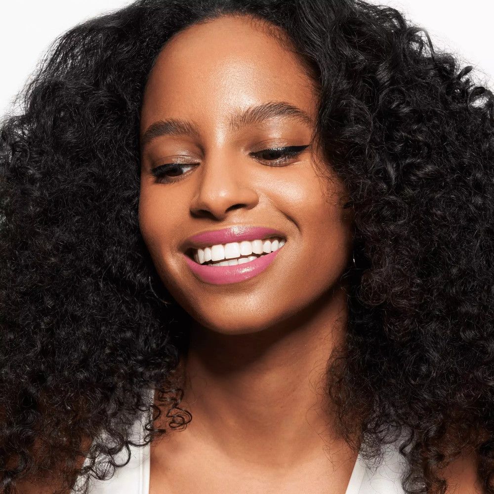 Porträt einer Frau mit rosa Lippen. Dunkle Locken, weißes Top. Lächelt.