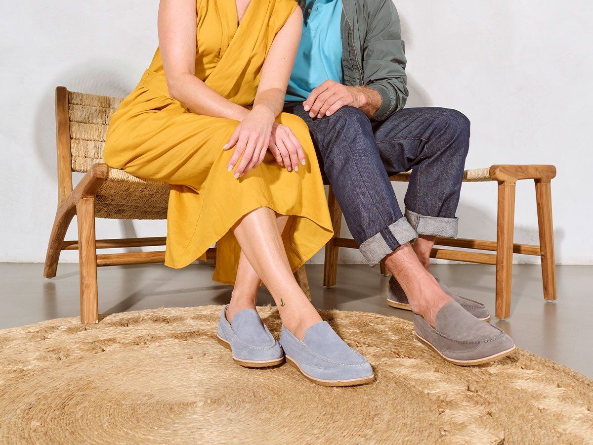 Personen sitzen auf einer Bank. Beide tragen Slipper. Frau: gelbes Kleid, blaue Slipper. Mann: Jeans, graue Slipper.