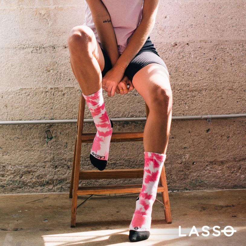Person sitzt auf Hocker. Trägt Kompressionssocken, Himbeerrot mit weißem Muster. Schwarze Zehen. Unten: LASSO-Logo.