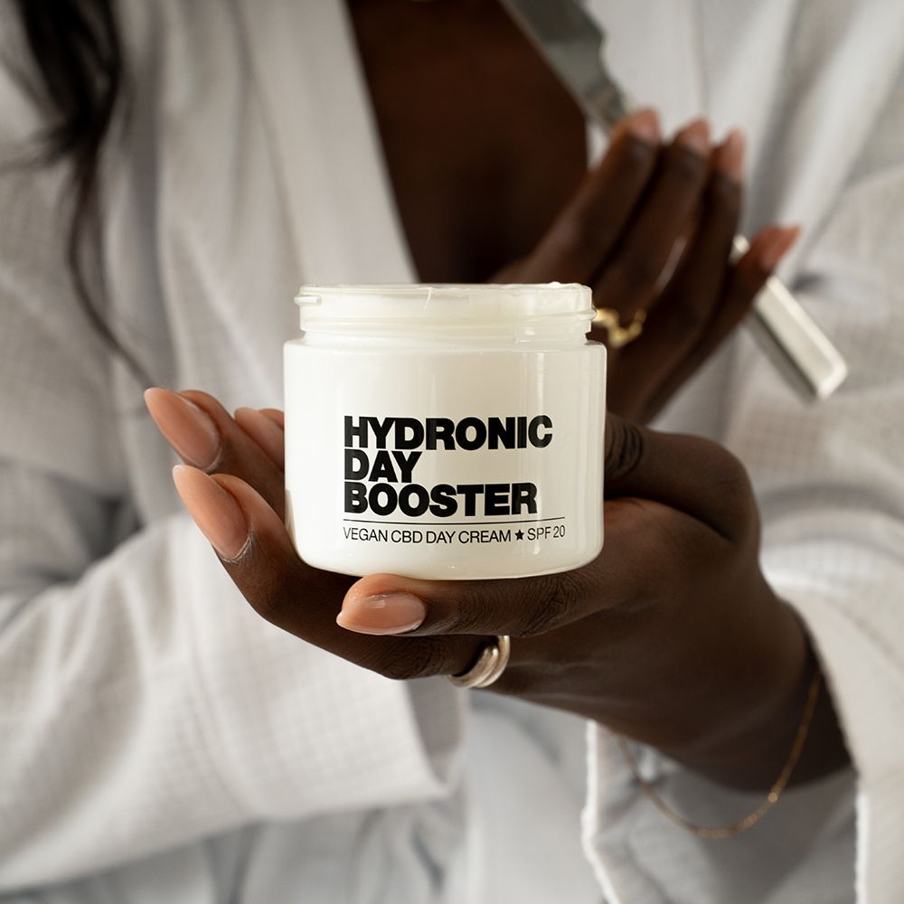 Weißes Tiegel mit Aufschrift: HYDRONIC DAY BOOSTER, Vegan CBD Day Cream + SPF 20, gehalten von einer Person in einem Bademantel.