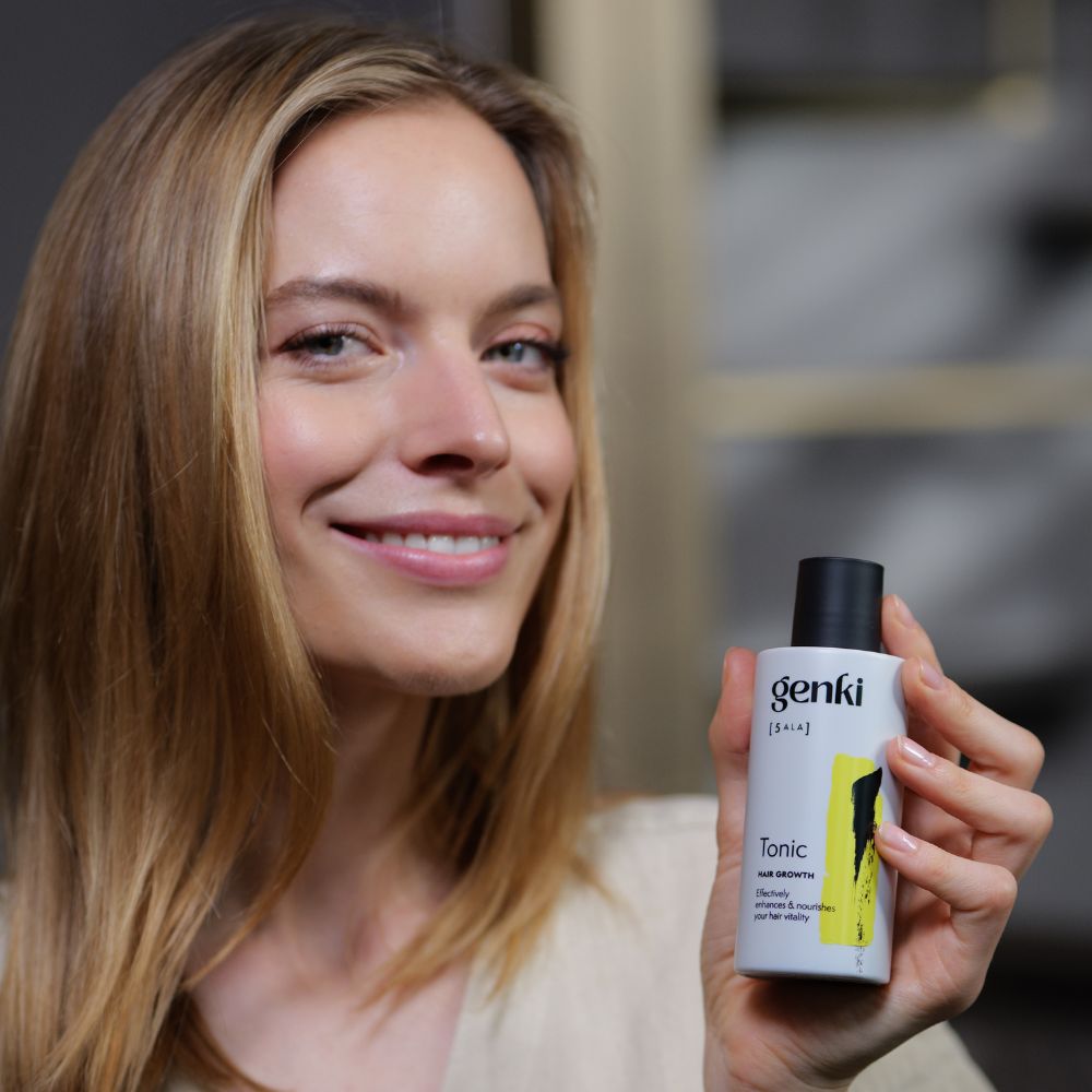 Frau hält Genki Tonic Flasche. Weiße Flasche mit schwarzem Deckel. Gelber Akzent. Schriftzug: Genki, Tonic.