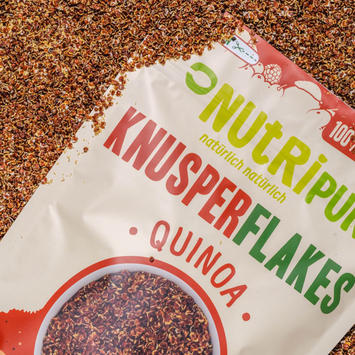 Nahaufnahme von NutriPur Knusperflakes Quinoa. Beutel teilweise im Bild. Text: Knusperflakes Quinoa.
