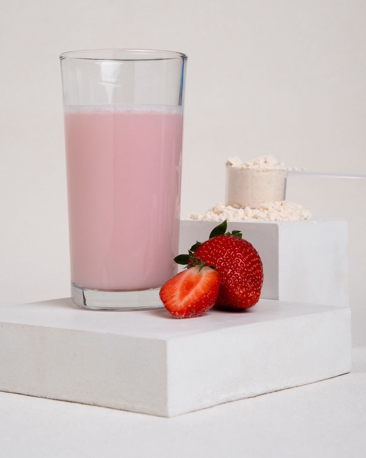 Glas mit rosa Flüssigkeit, Erdbeeren und Messlöffel mit Proteinpulver auf weißem Untergrund.
