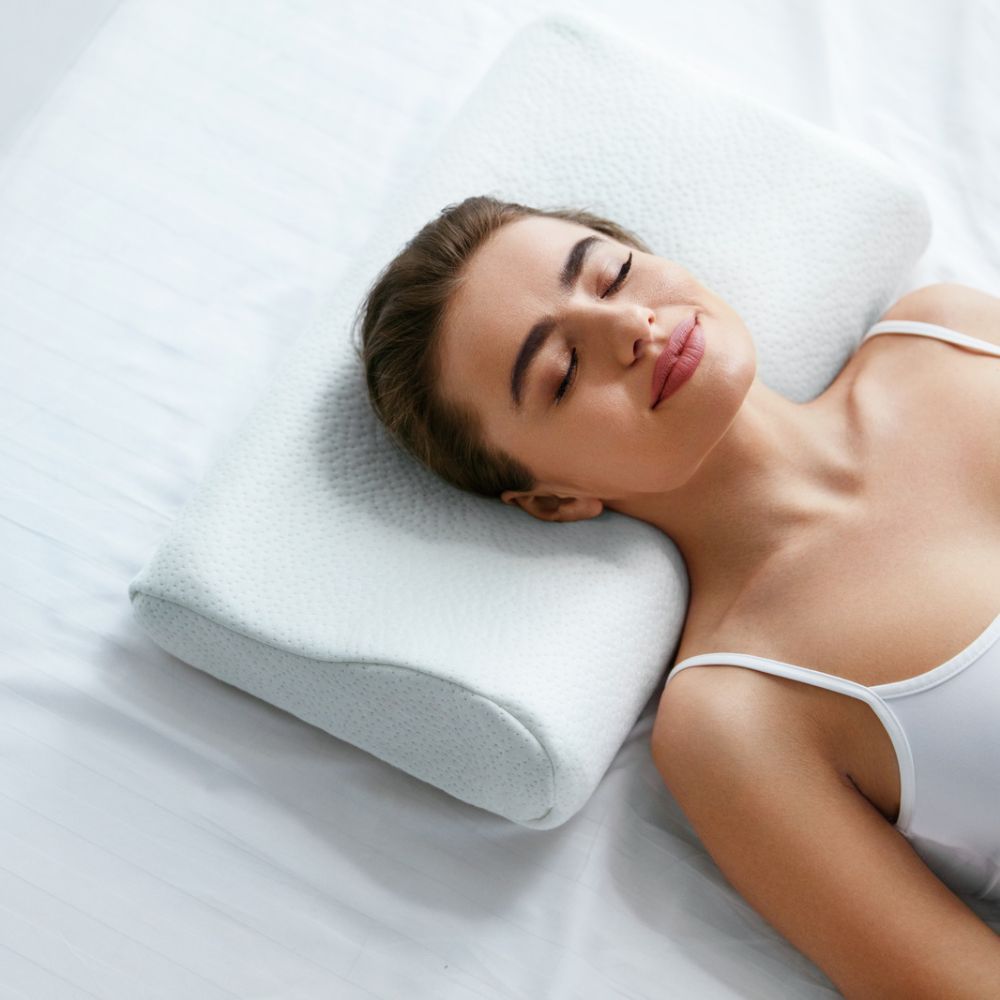 Femme allongée sur le dos, tête sur un oreiller cervical blanc. Yeux fermés. Haut blanc.