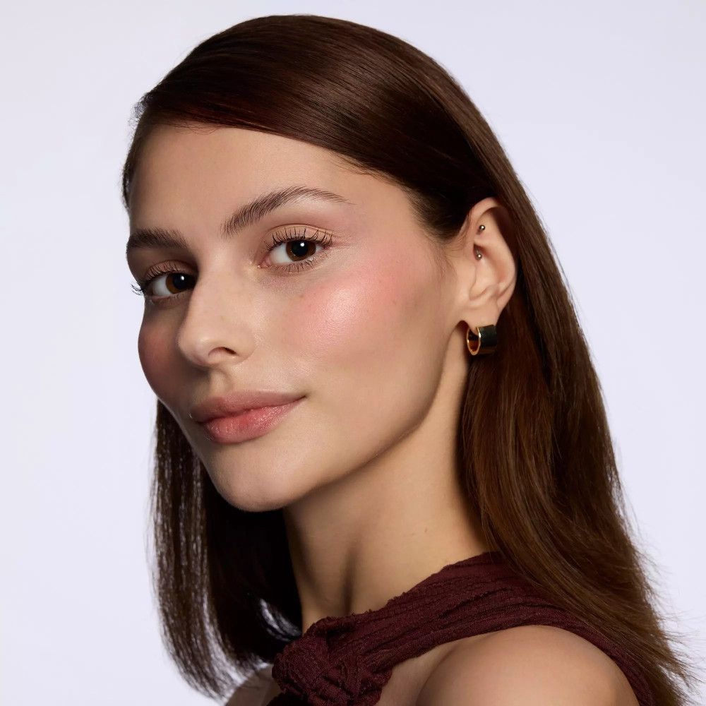 Porträt einer Frau mit Rouge auf den Wangen. Sie trägt Ohrringe. Braune Haare. Heller Hintergrund.