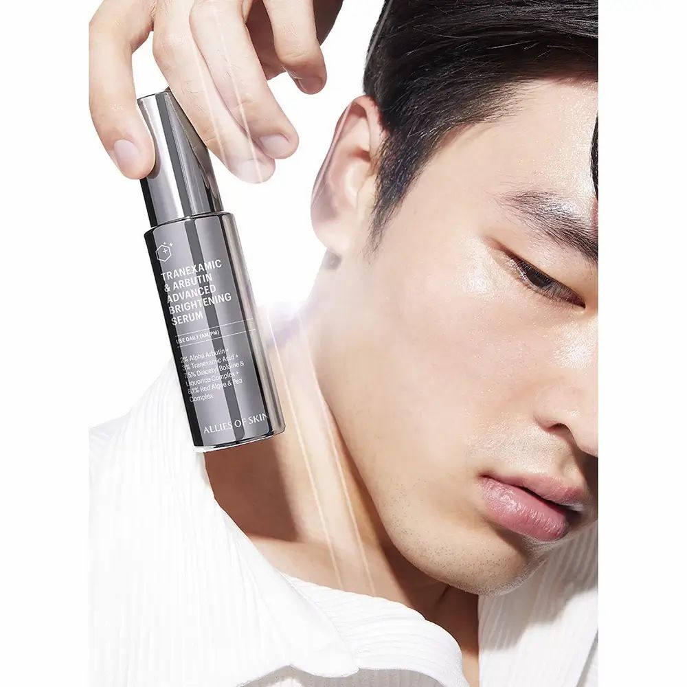 Mann hält eine dunkle Flasche mit Sprühkopf. Beschriftung: Tranexamsäure & Arbutin Advanced Brightening Serum. Markenname.