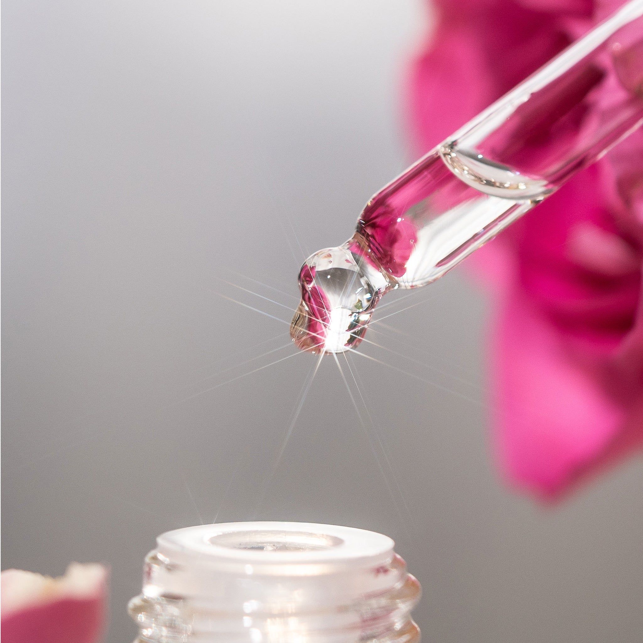Nahaufnahme einer Pipette mit Tropfen. Flüssigkeit tropft in eine Flasche. Hintergrund: rosa Blume.