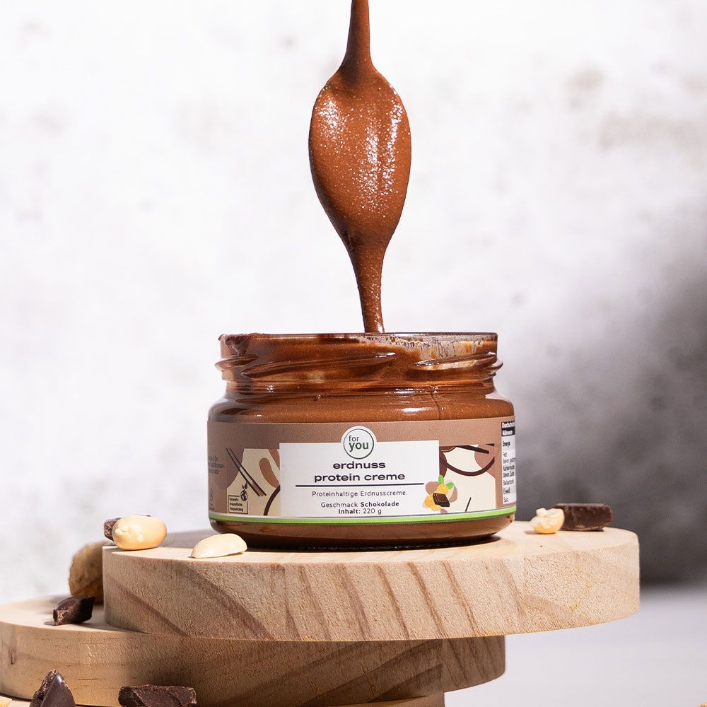Glas mit brauner Creme, Löffel mit Creme darüber. Auf Holzscheiben platziert, umgeben von Erdnüssen und Schokoladenstücken.